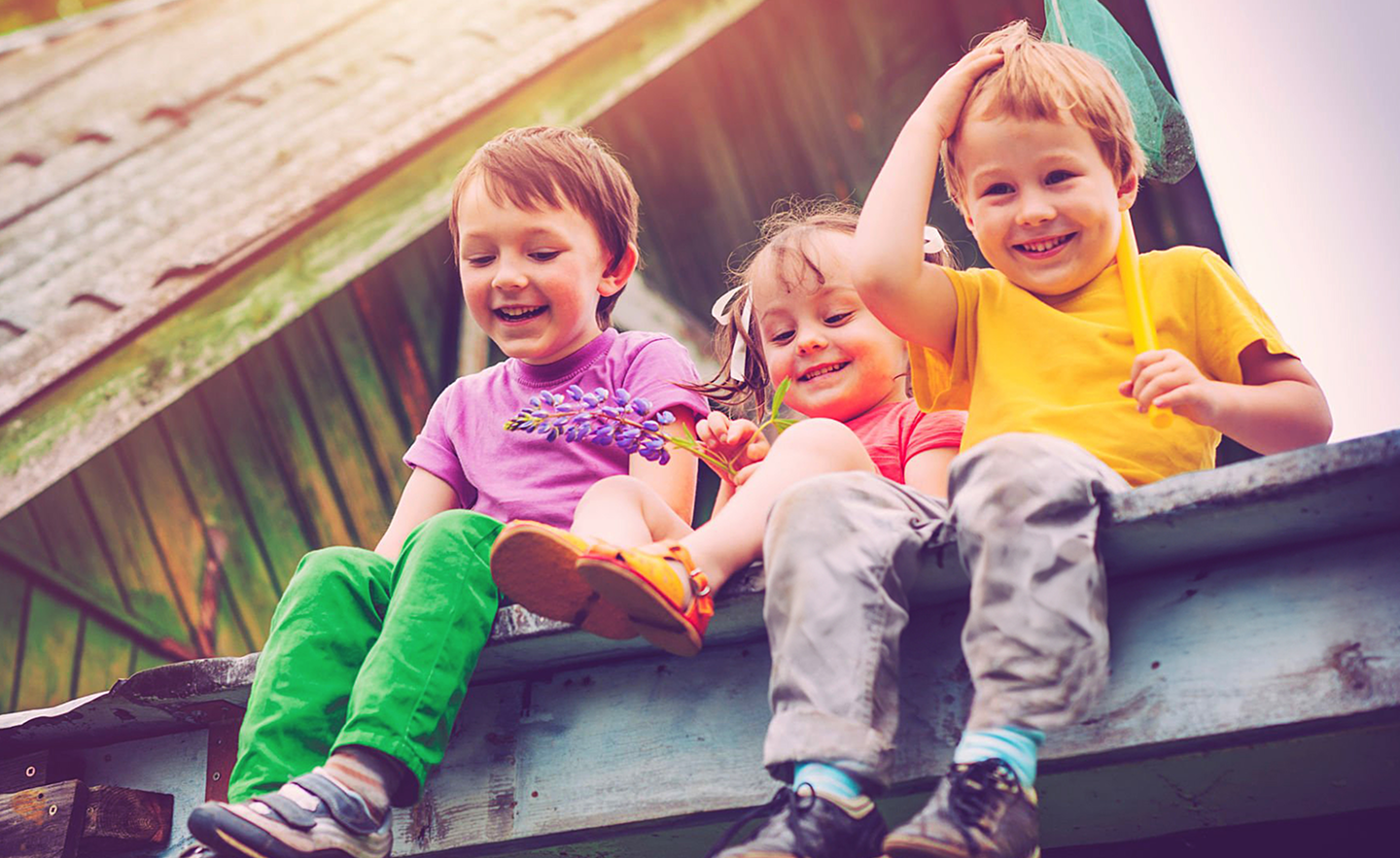 Drei Kinder sitzen lachend auf einem Dachrand und spielen im Freien